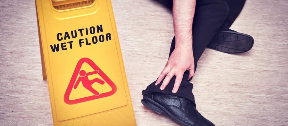 Person sitting on the floor holding their ankle near a yellow "Caution Wet Floor" sign on a tiled surface.