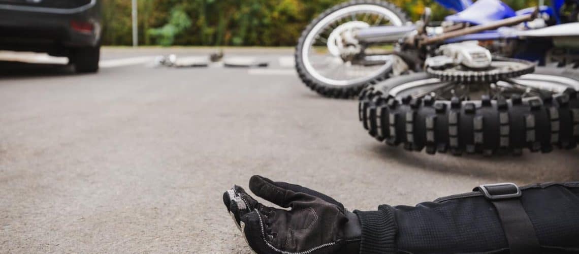 A motorcyclist wearing black gloves lies on the ground near a fallen motorcycle on a road with blurred greenery in the background.