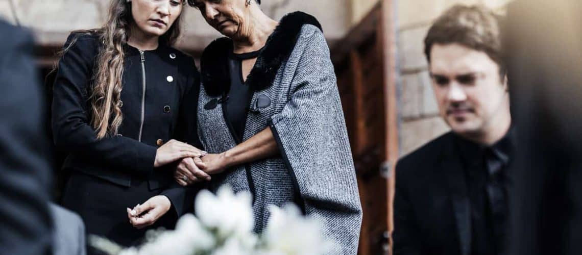 People standing together in sombre clothing, holding hands, with a blurred floral arrangement in the foreground.