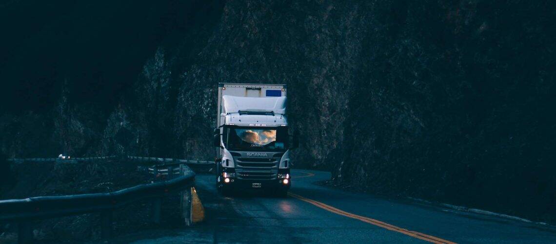 Truck driving on a winding mountain road at dusk, surrounded by steep rock faces and dimly lit by the remaining daylight.