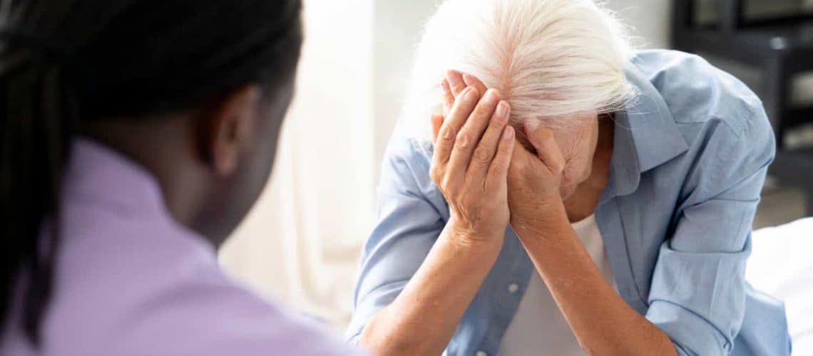 An elderly person sitting with their hands on their face, appears distressed. Another person facing them listens intently.