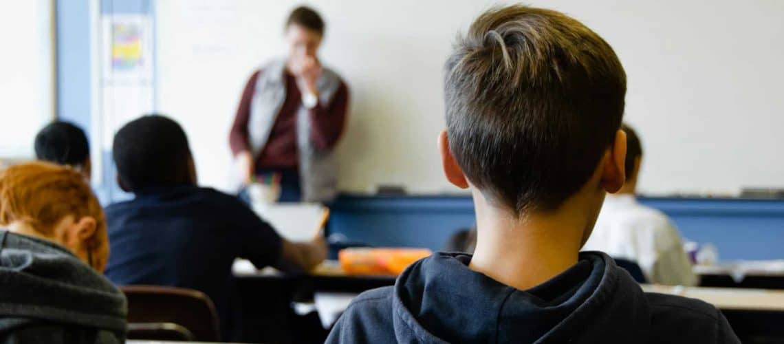 Students seated at desks facing a teacher in front of a whiteboard in a classroom setting.