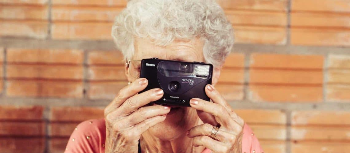 An elderly woman holds a Kodak camera up to her face, standing against a brick wall.