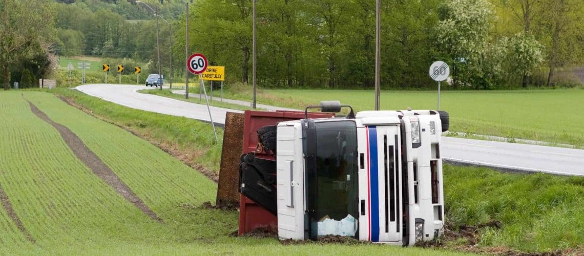 A truck is overturned on a grassy field beside a road. There's a speed limit sign and a curve warning ahead.