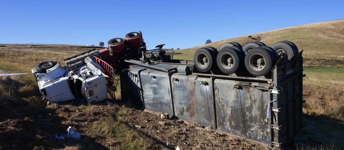 Overturned semi-truck on its side in a grassy area, showing the trailer with six visible wheels against a clear blue sky.