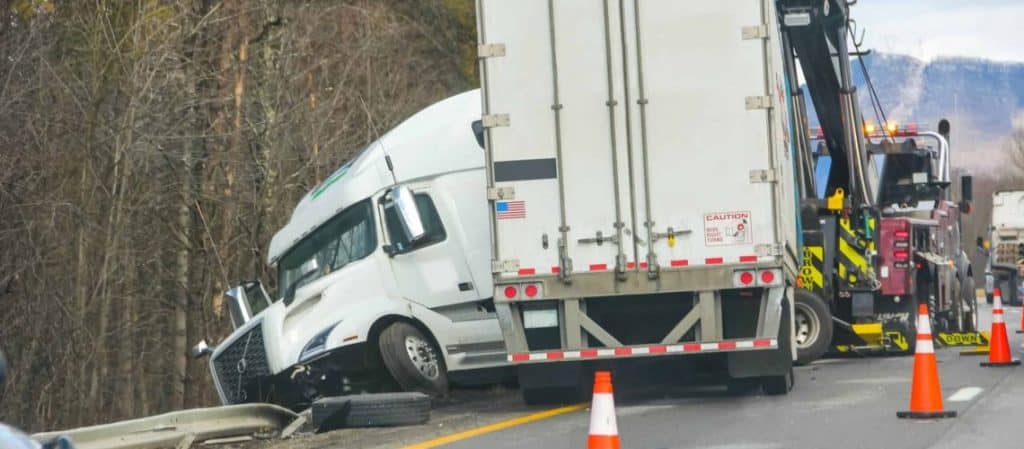 A large white semi-truck is involved in an accident on the highway, partially off the road. Traffic cones and a recovery vehicle are present.