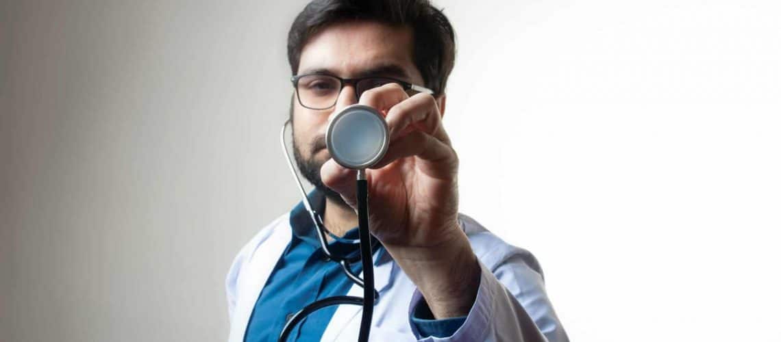 A person in a white coat holds a stethoscope towards the camera against a plain background.