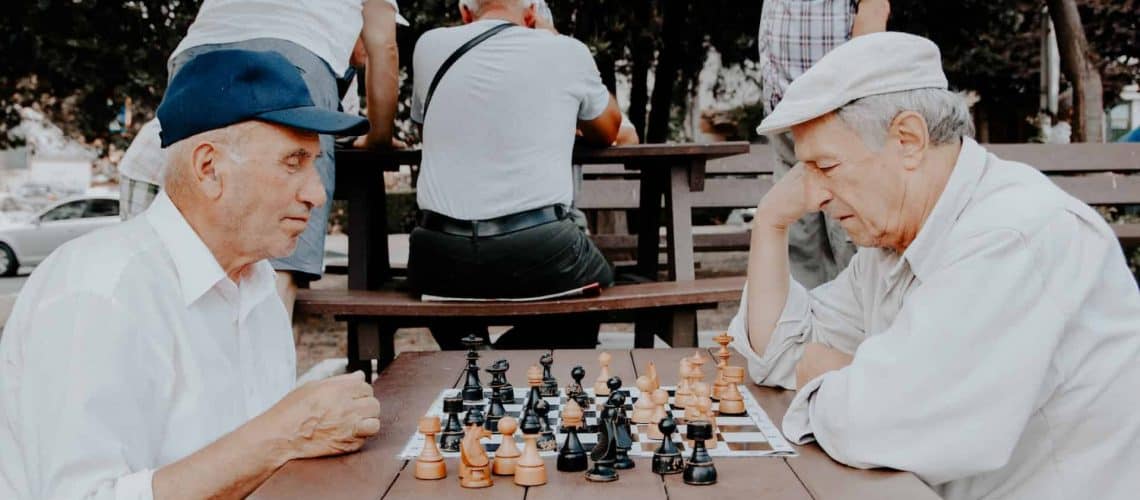 Two elderly men in caps play chess at an outdoor table. Other individuals are seated on benches in the background.