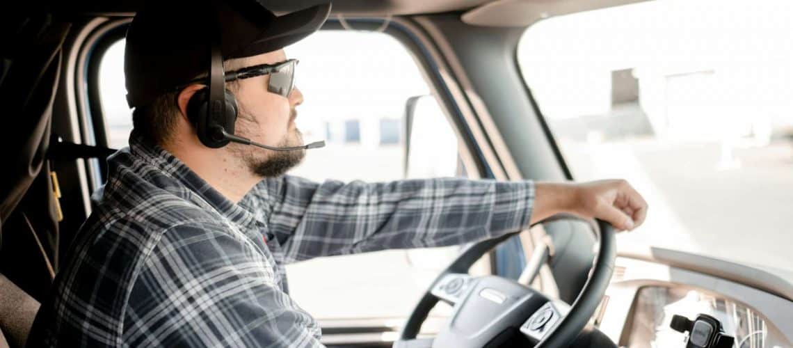 A man wearing a cap, sunglasses, and a headset is driving a truck, looking focused. He is wearing a plaid shirt and holding the steering wheel with one hand.