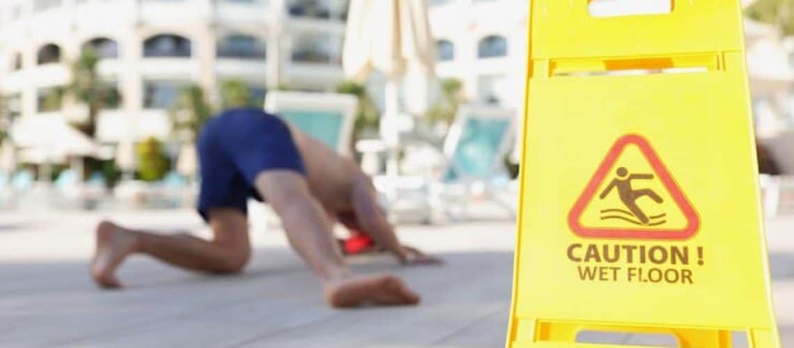 Yellow "Caution! Wet Floor" sign in focus, with a person on the ground in the blurred background near a pool.